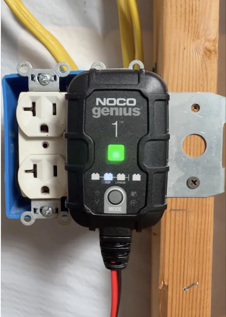 A NOCO Genius 1 battery tender mounted on a garage wall, green light on, maintaining a motorcycle battery