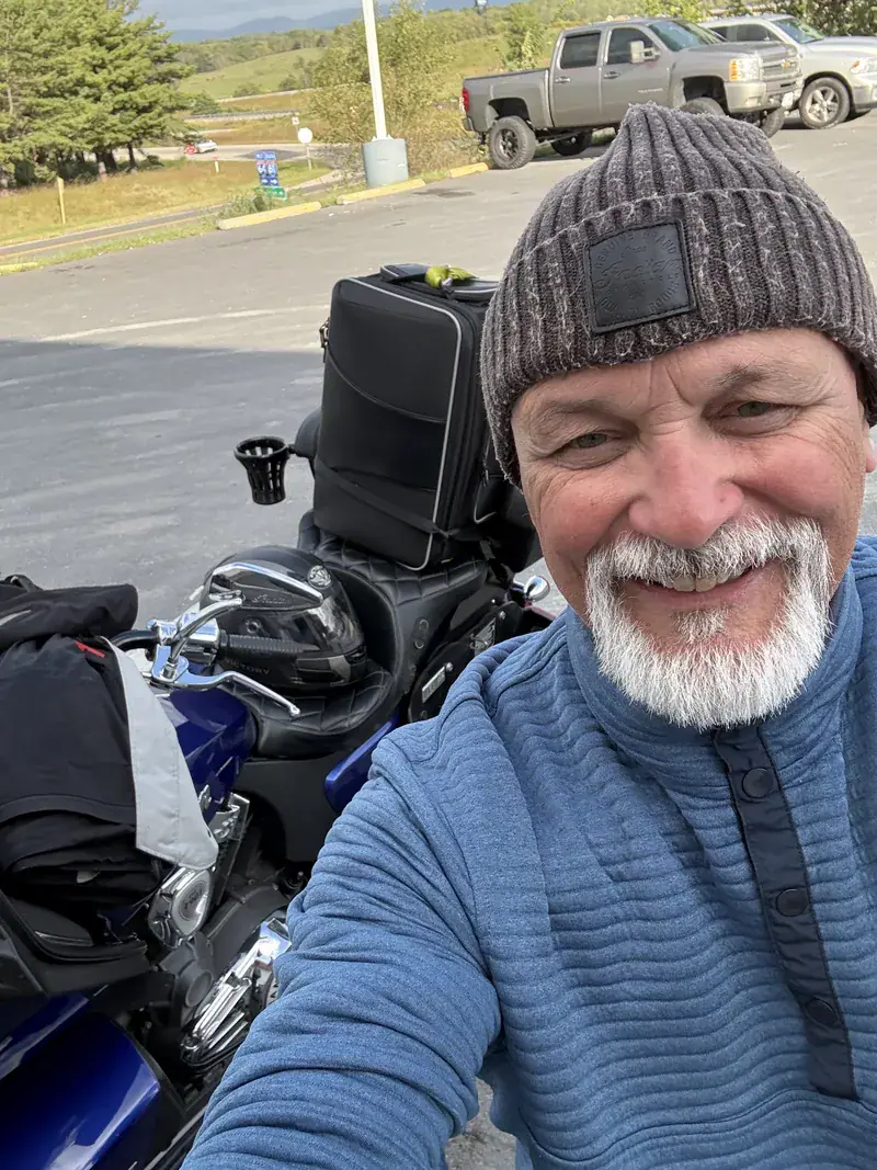 Rider in a knit beanie smiling next to his loaded blue touring motorcycle at a stop in the Virginia mountains